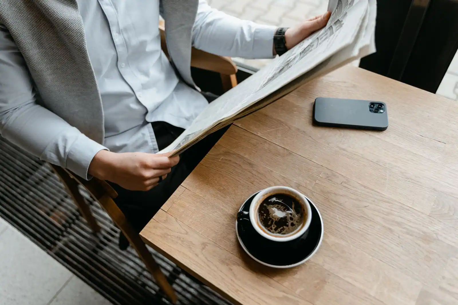 Personne lisant un journal papier attablée dans un café, une tasse et un smartphone posés sur la table