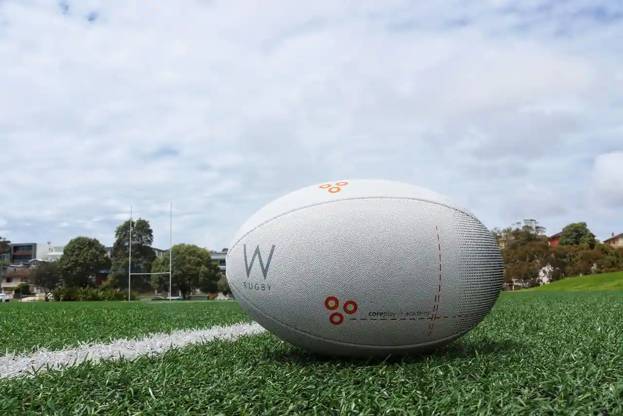 Ballon de rugby blanc posé sur la pelouse d'un terrain, poteaux en H visibles à l'arrière-plan