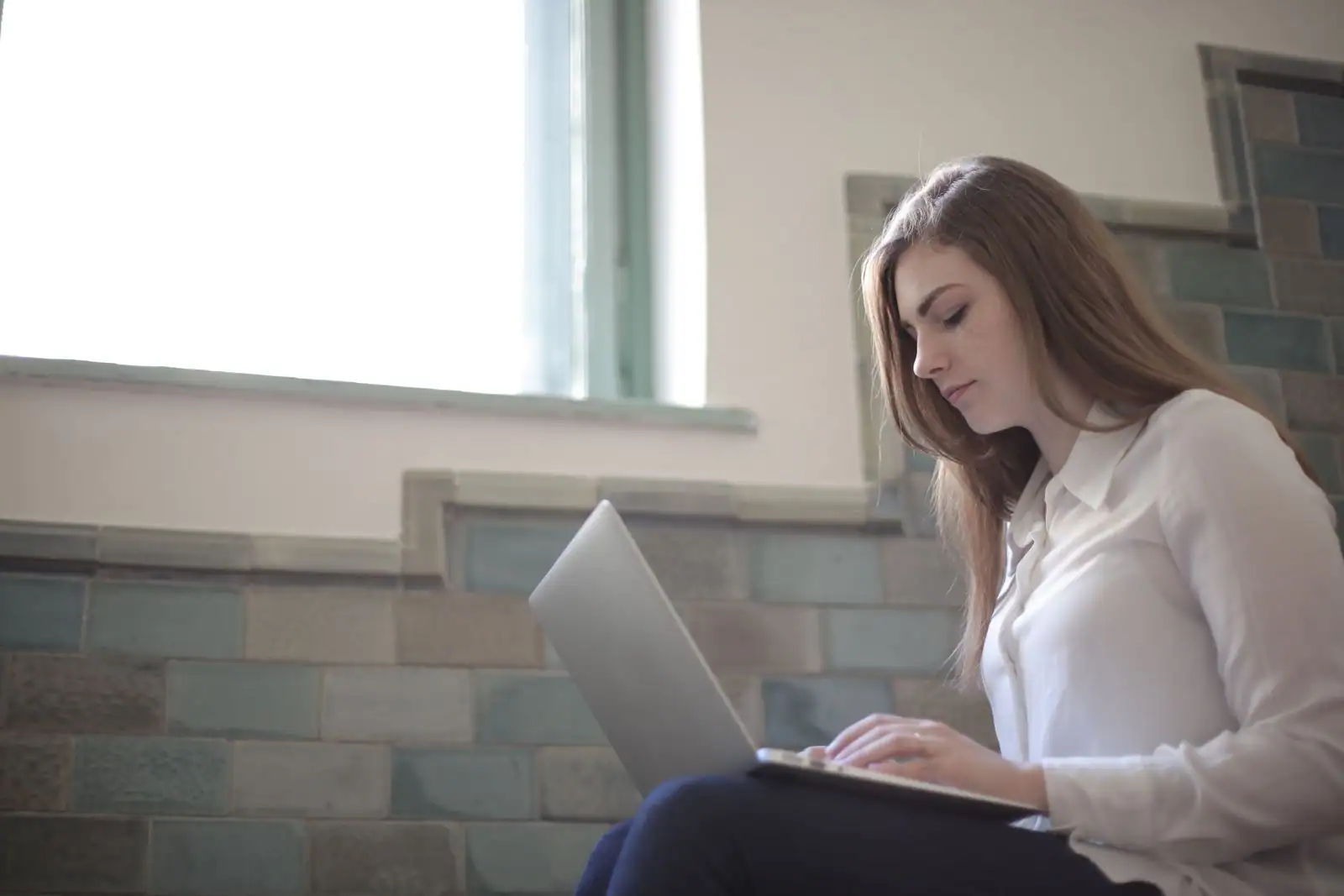 Femme en chemise blanche tapant sur son ordinateur portable, assise près d'une fenêtre