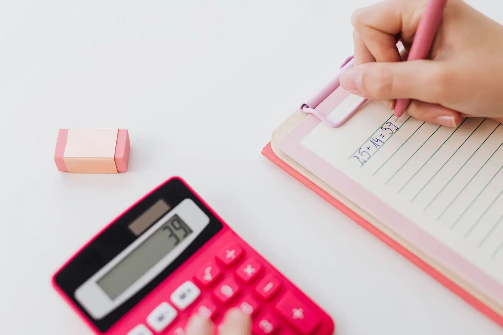 Main écrivant une opération mathématique sur un carnet, à côté d'une calculatrice rose.