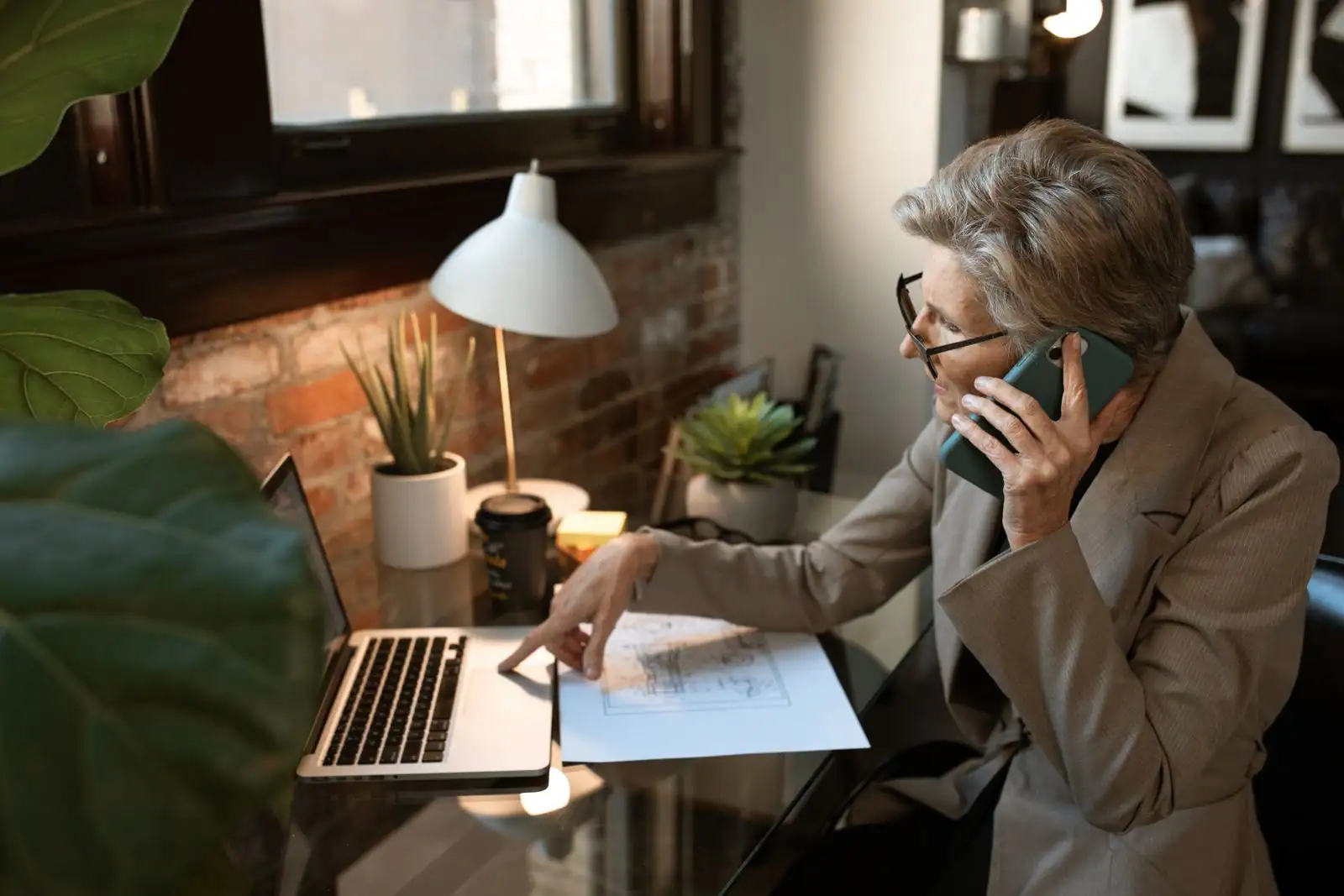 Une personne tient un smartphone à l'oreille devant un ordinateur portable ouvert, à son bureau.
