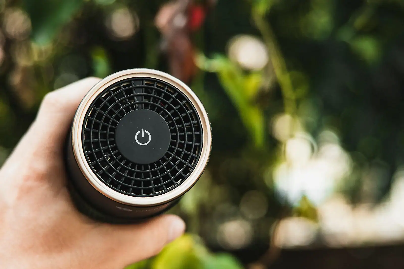 Main tenant une petite enceinte Bluetooth portable cylindrique, vue de dessus, sur fond de verdure flou.