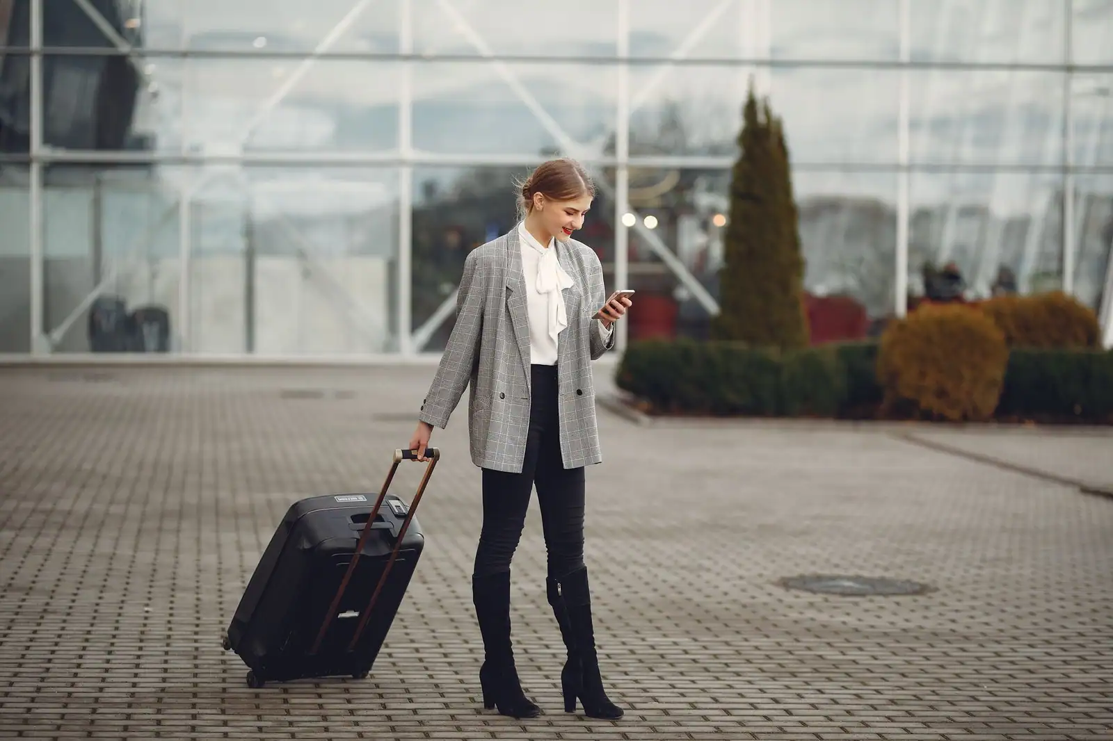 Voyageuse avec une valise à roulettes consultant son smartphone devant la façade vitrée d'un aéroport.