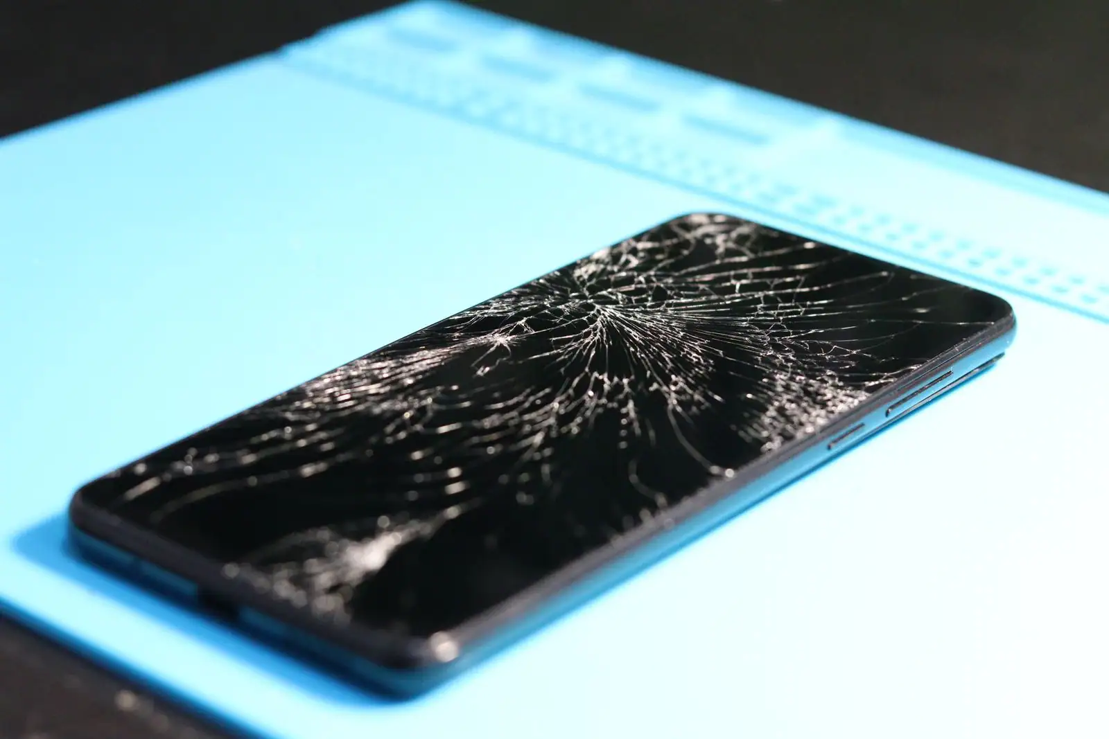 Smartphone à l'écran très fissuré posé sur un tapis de réparation bleu, la vitre brisée bien visible