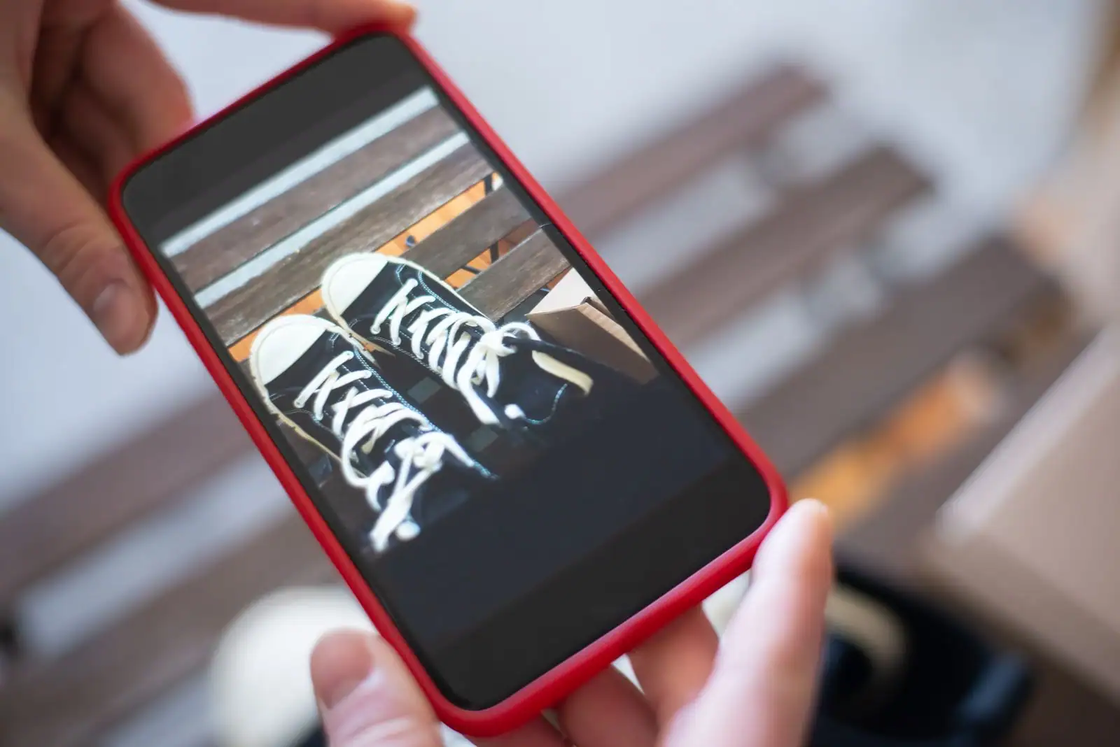 Smartphone moderne dans une coque rouge, tenu à deux mains, écran allumé affichant une photo de baskets.