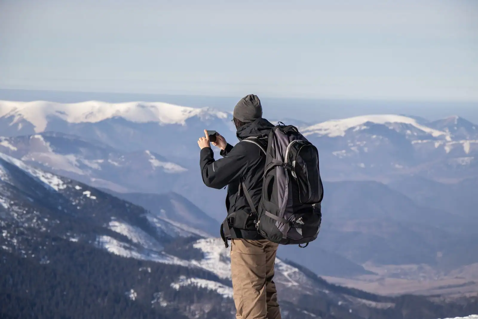 Randonneur avec un grand sac à dos, smartphone en main, face à un massif de montagnes enneigées.