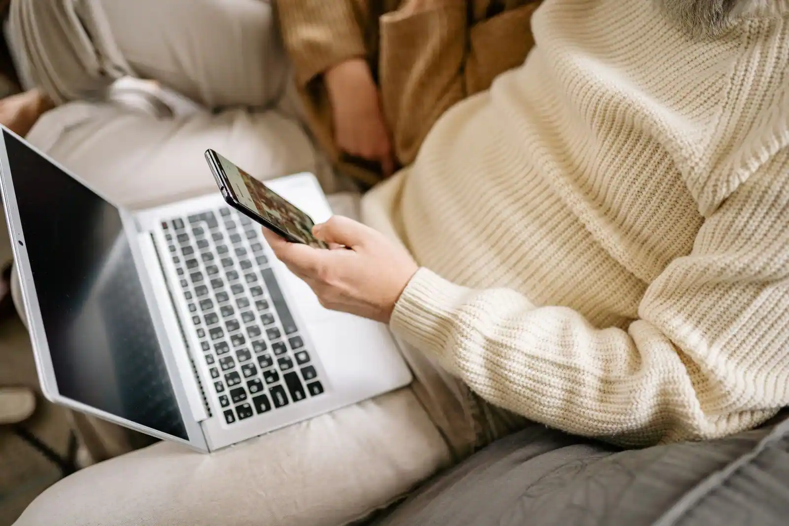 Personne tenant un smartphone, un ordinateur portable ouvert posé sur les genoux