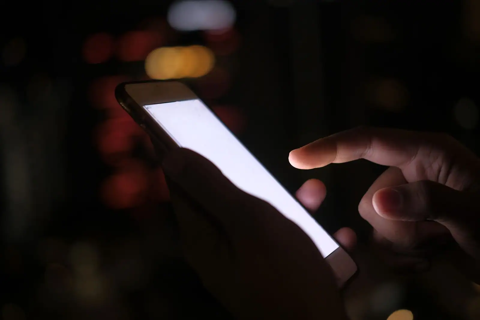 Main tenant un smartphone à l'écran allumé, lumières de la ville floutées en arrière-plan.