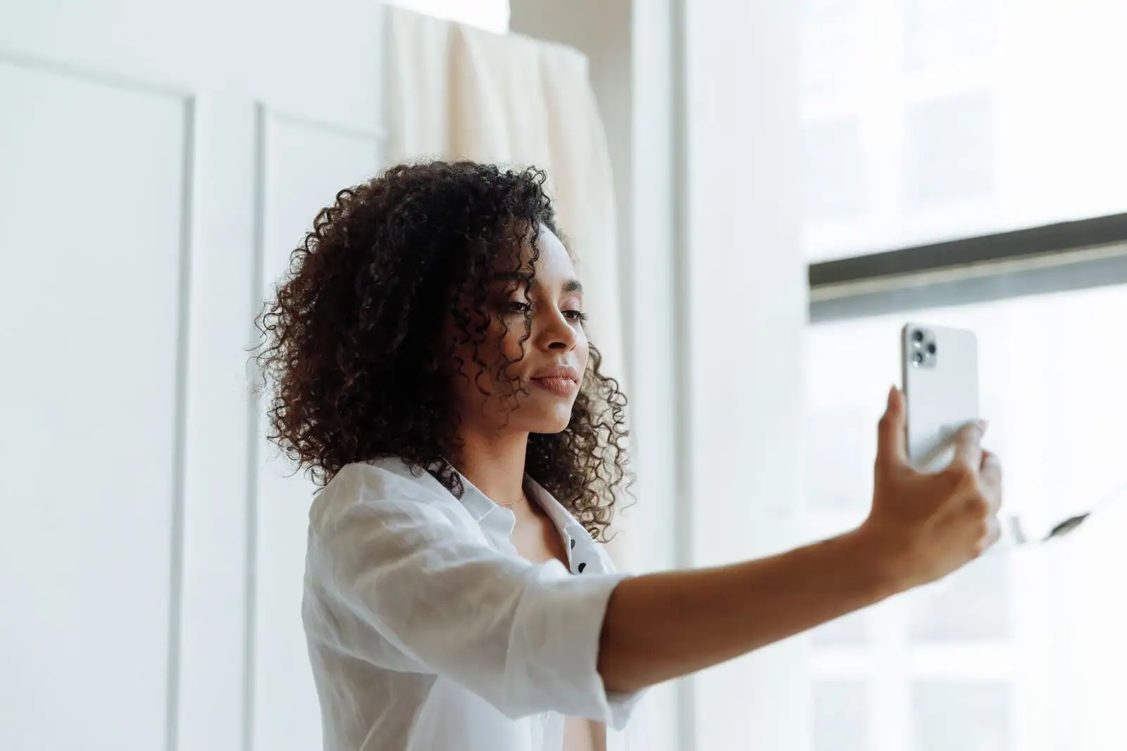 Jeune femme tenant son smartphone à bout de bras pour se photographier devant un fond clair.