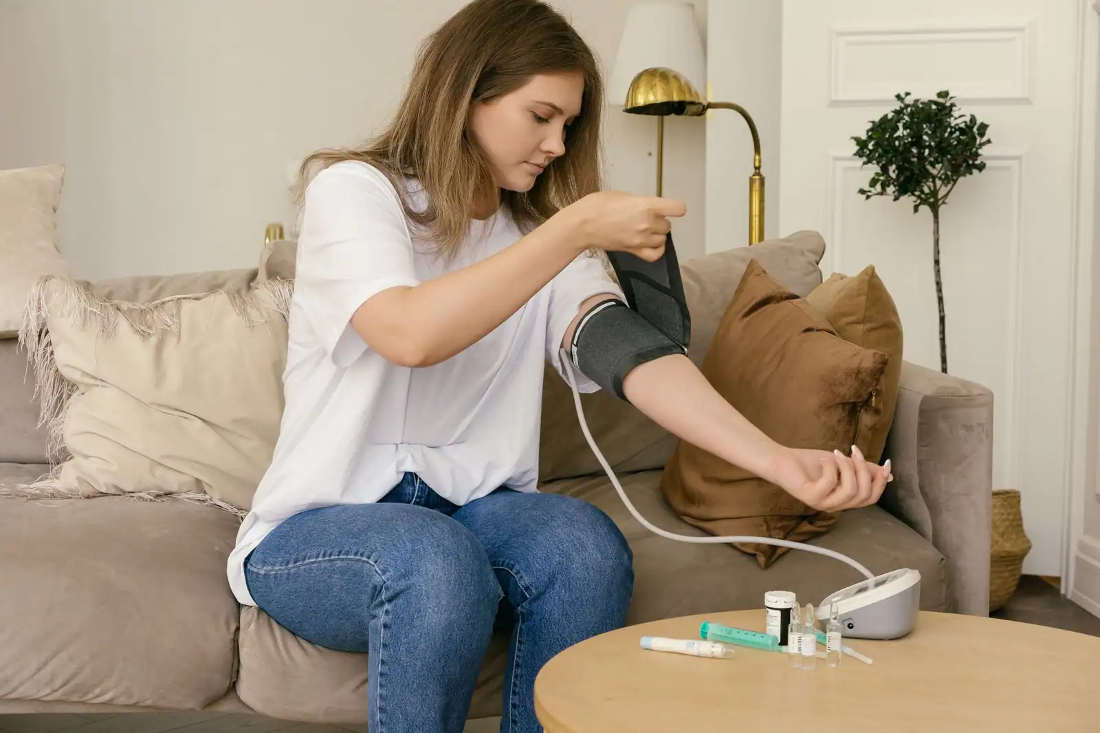 Jeune femme installant le brassard d'un tensiomètre sur son bras, assise sur un canapé, pour une mesure à domicile