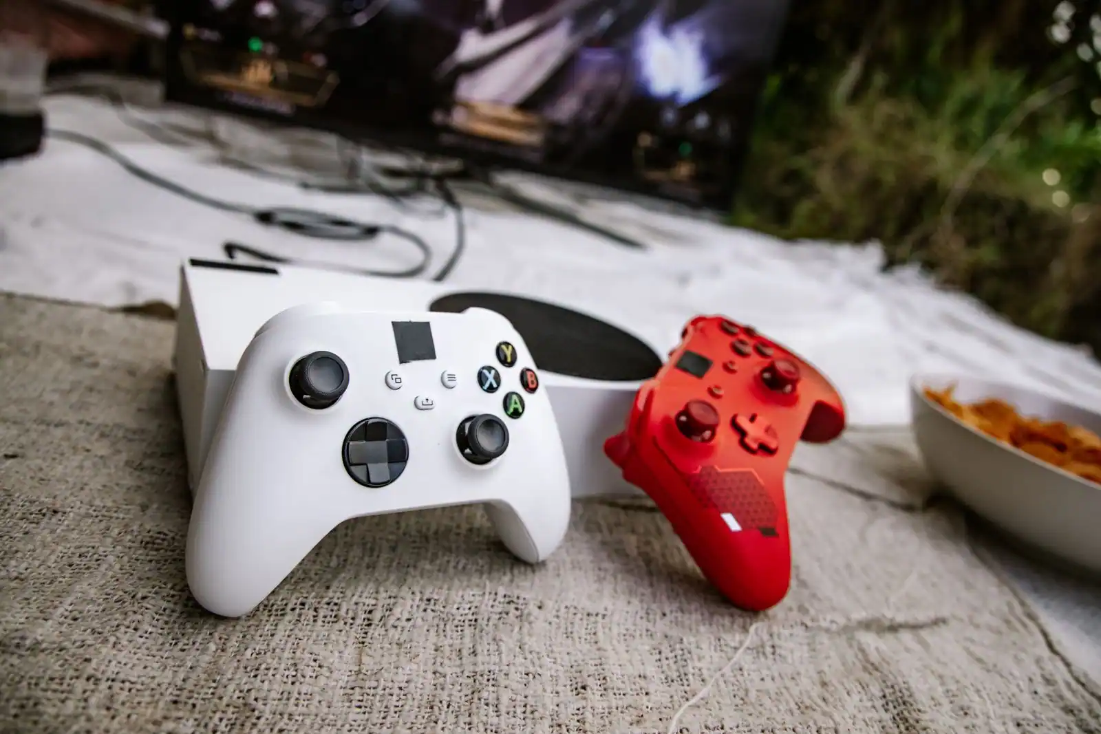 Deux manettes Xbox, une blanche et une rouge, posées devant une console blanche et un écran allumé.