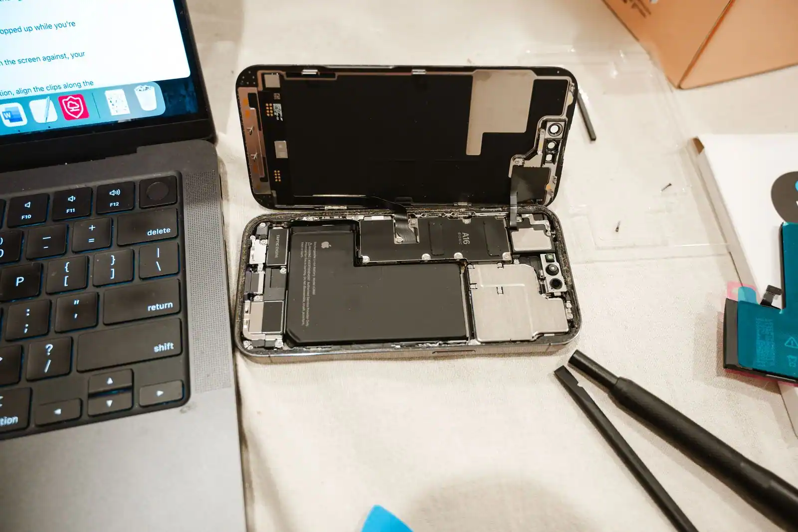 Un iPhone ouvert sur un plan de travail, batterie d'origine visible, module neuf et outils de démontage à côté.