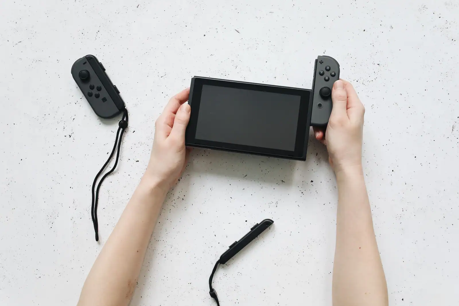 Deux mains tiennent une console Nintendo Switch écran éteint, une manette latérale détachée posée à côté