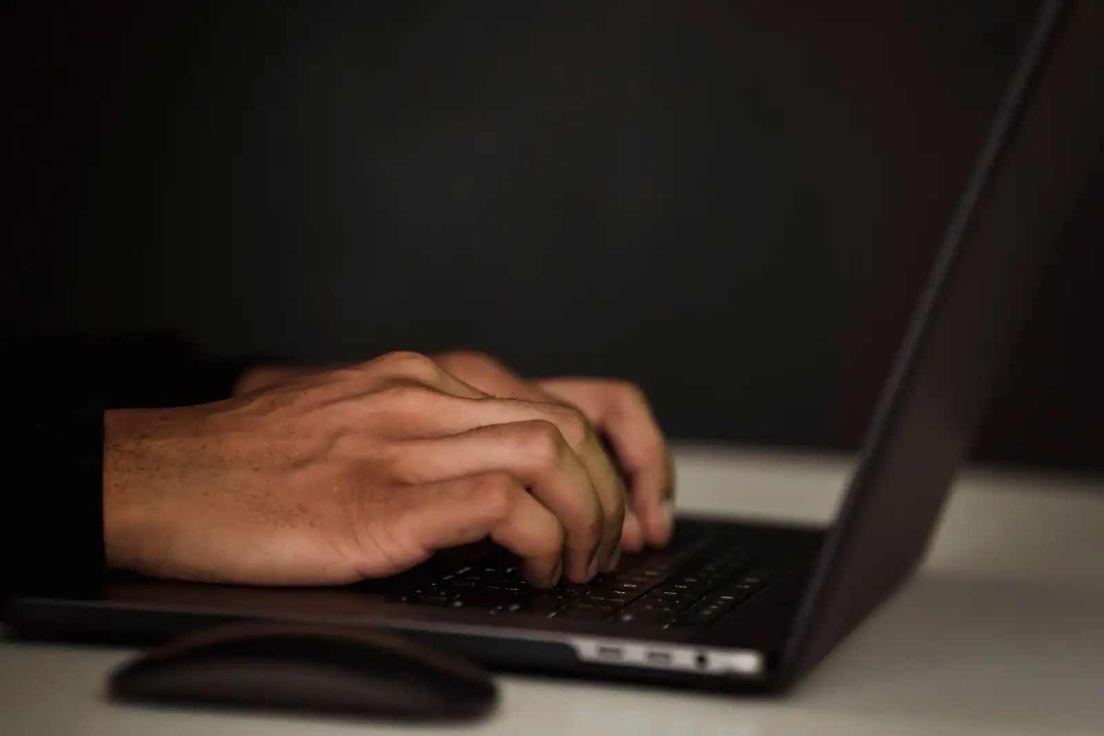 Mains d’une personne en train de taper sur le clavier d’un ordinateur portable, pièce sombre