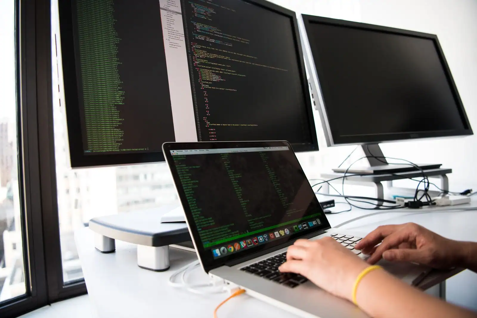 Mains sur le clavier d'un ordinateur portable, devant deux écrans affichant des lignes de code