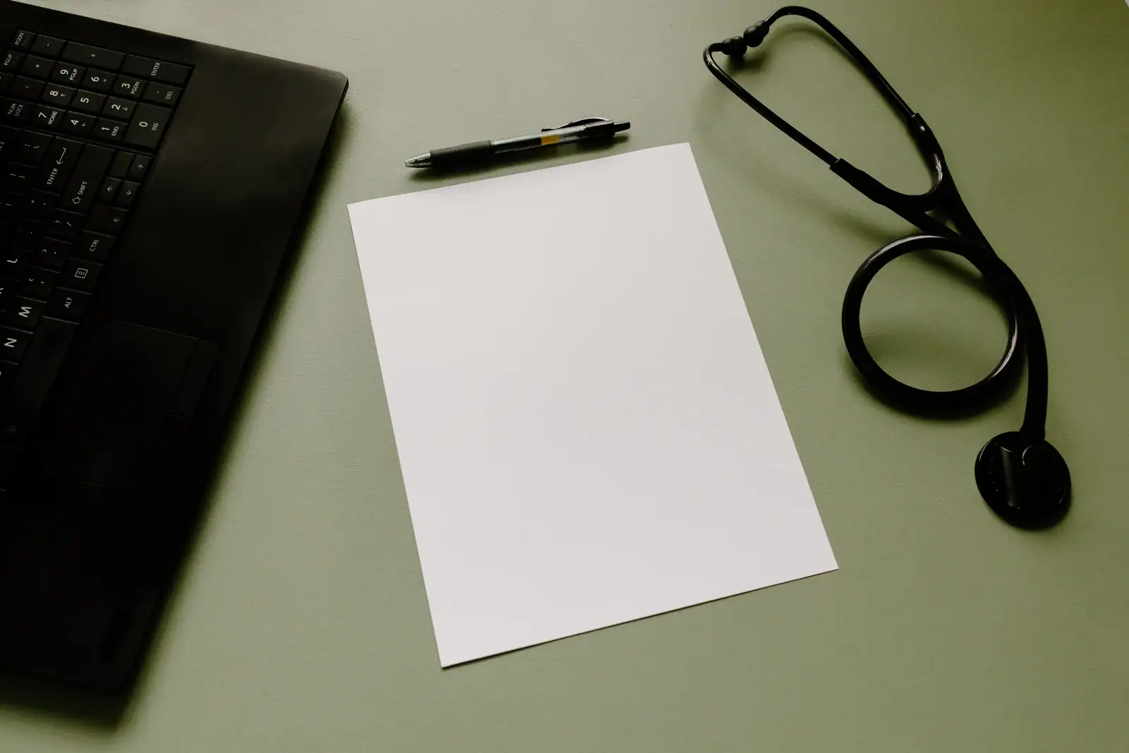 Ordinateur portable, feuille vierge et stéthoscope posés sur un plan de travail vert pâle