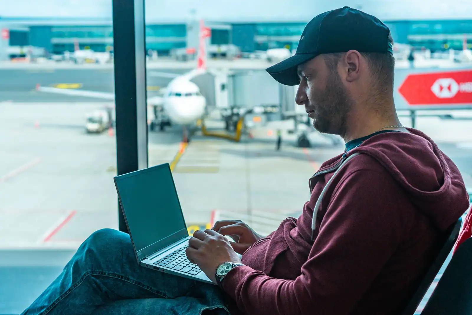 Homme dans un terminal d'aéroport, ordinateur portable sur les genoux, avion stationné derrière la vitre.