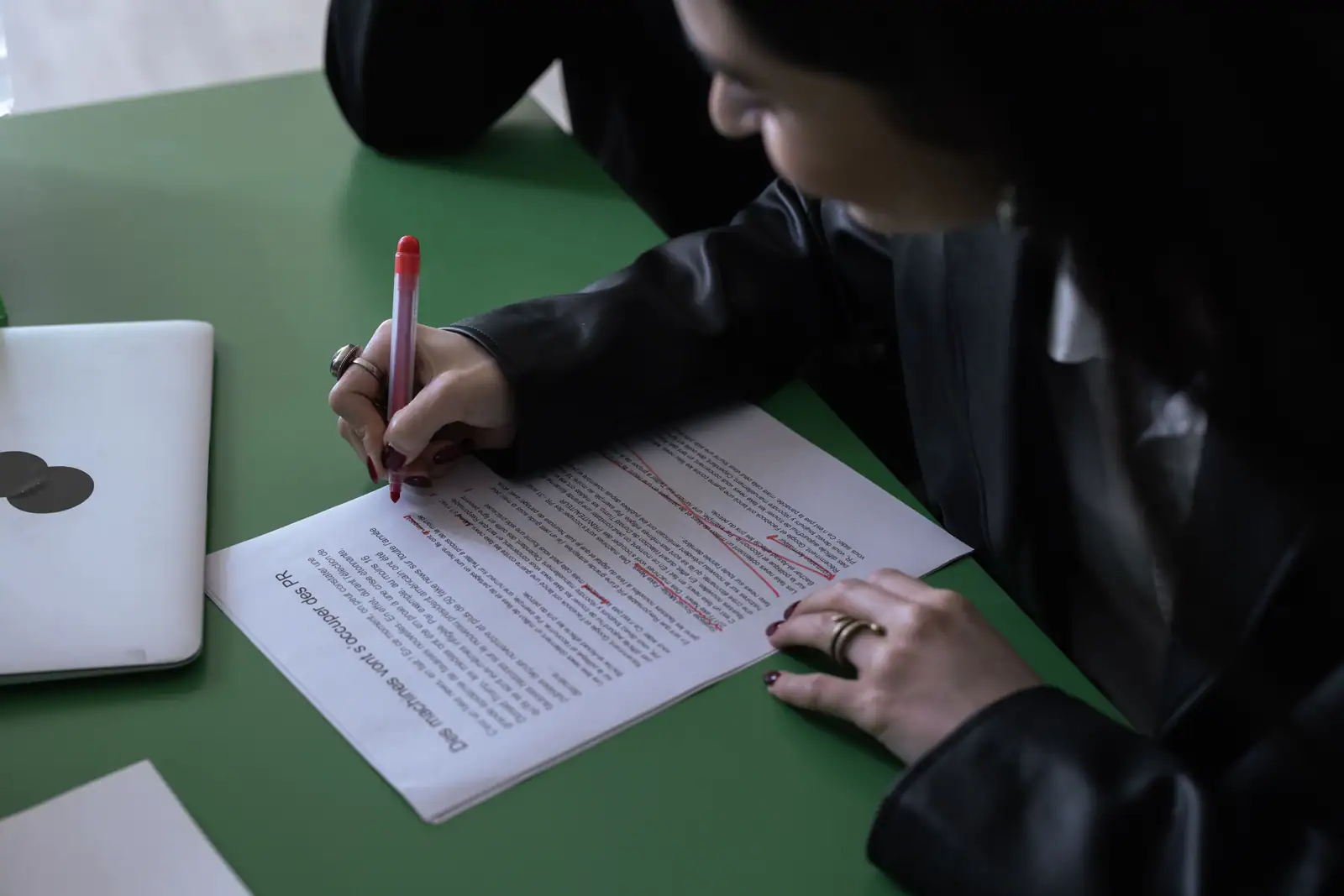 Une femme annote au stylo rouge un texte imprimé posé sur une table verte, près d'un ordinateur portable fermé