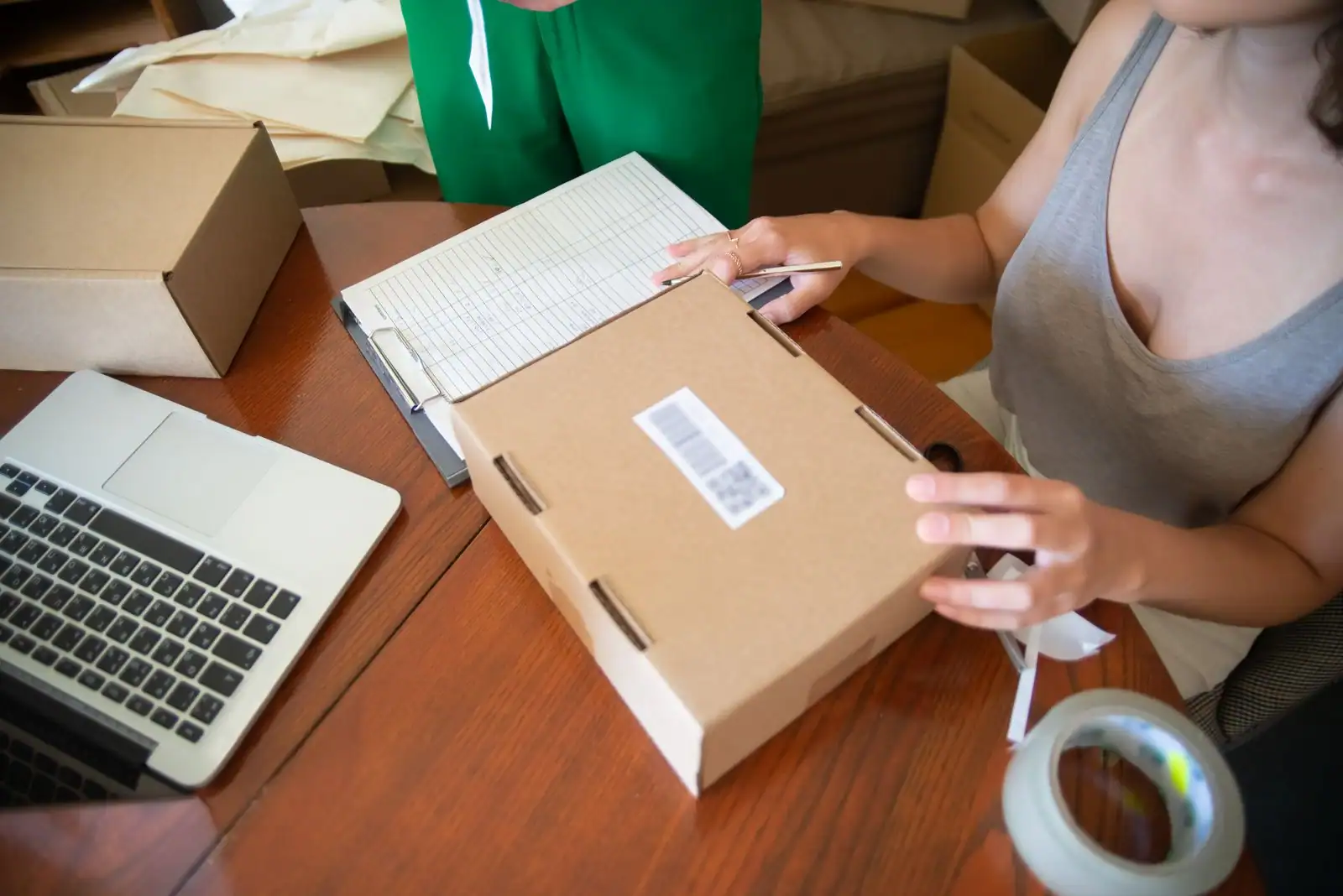 Vendeuse en ligne scellant un colis étiqueté sur une table de travail, ordinateur portable posé à côté