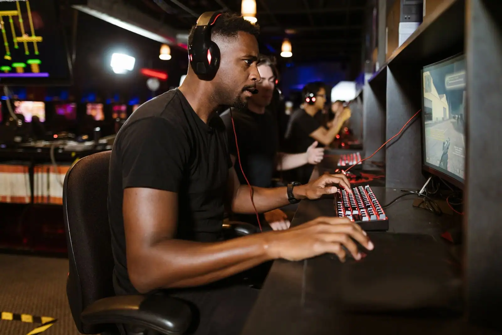 Joueur au casque-micro devant son écran, une main sur un clavier rétroéclairé, dans une salle de jeu sombre.