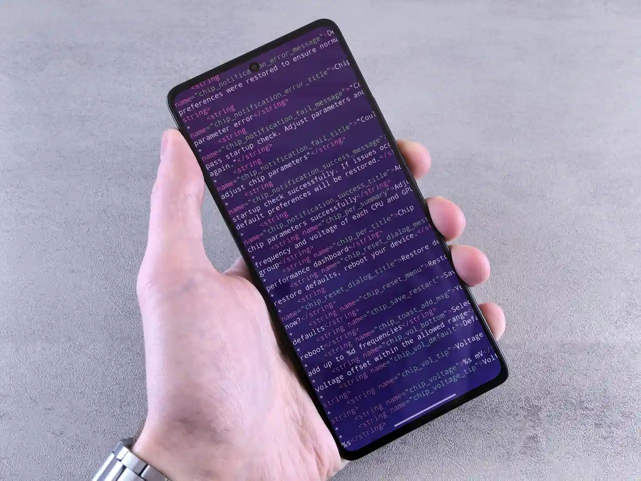 Main tenant un smartphone dont l'écran affiche des paramètres système en texte, sur un plan de travail gris.
