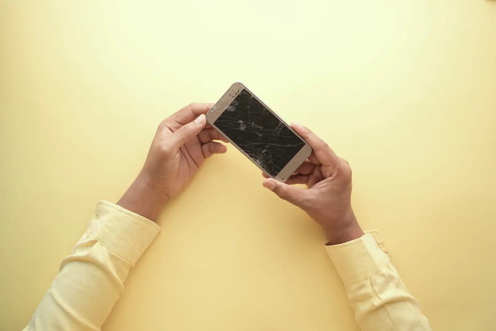 Deux mains tenant un smartphone à l’écran fissuré, vu de dessus sur un fond jaune uni