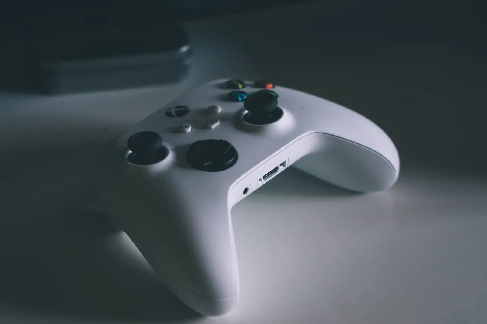 Manette Xbox blanche posée sur une surface claire dans une lumière basse, la console floue en arrière-plan.